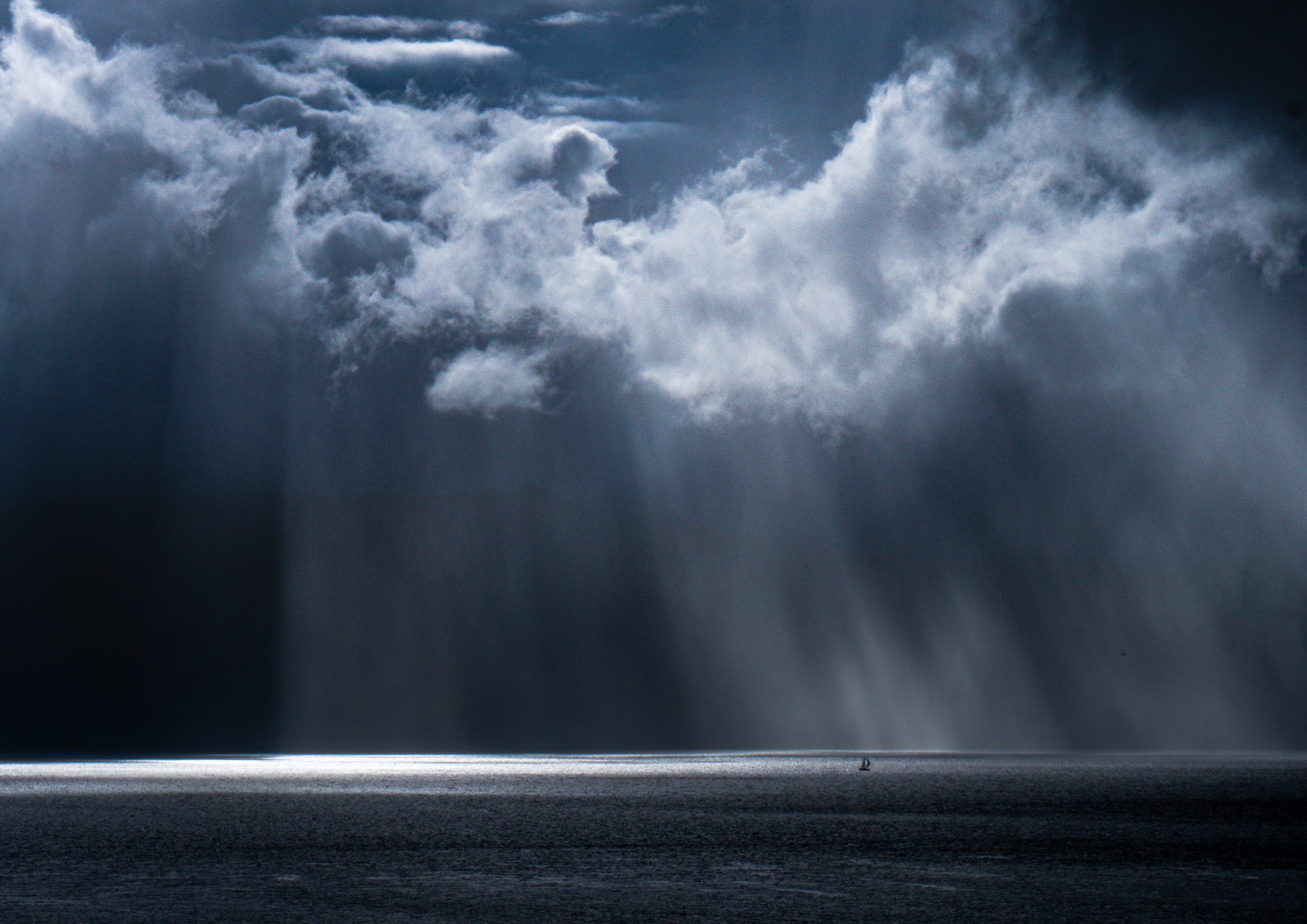 A solitary sailboat beneath an immense sky with shafts of light reaching the water